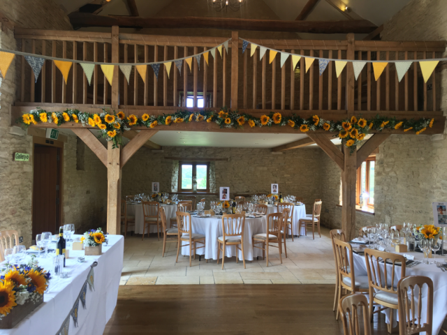 Balcony and table displays including Sunflower, Gypsophila and touches of bright blue Delphinium at The Kingscote Barn. Floral design by Cotswold Blooms, wedding florist based in Cheltenham.