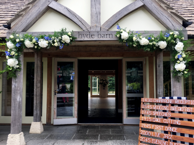 Hyde Barn entrance dressed with white and blue Hydrangea, Delphinium and Rose displays. Floral design by Cotswold Blooms, wedding florist based in Cheltenham.