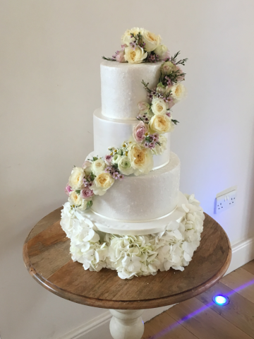 Roses, Wax Flower and Ranunculus on top and elegantly wrapped around the wedding cake with a Hydrangea base. Floral design by Cotswold Blooms, wedding florist based in Cheltenham.