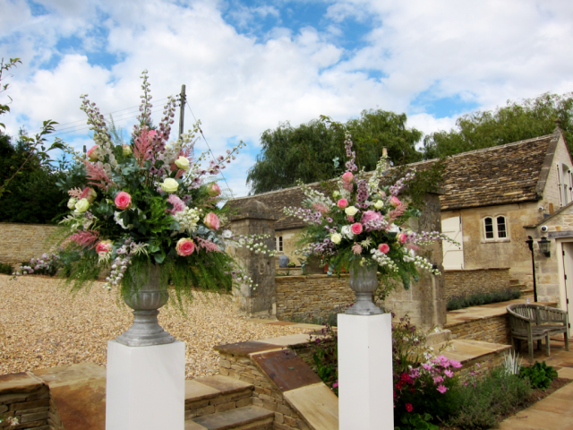 Stone urn displays with Delphinium, Roses, Peonies and Thlaspi for an outdoor ceremony. Floral design by Cotswold Blooms, wedding florist based in Cheltenham.