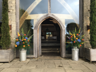 Bright orange and blue milk churn including Gladioli, Dahlia and Roses outside Cripps Barn. Floral design by Cotswold Blooms, wedding florist based in Cheltenham.