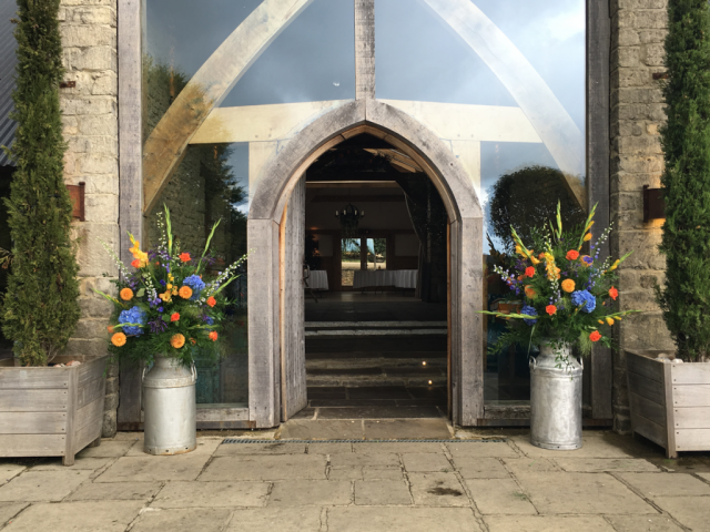 Bright orange and blue milk churn including Gladioli, Dahlia and Roses outside Cripps Barn. Floral design by Cotswold Blooms, wedding florist based in Cheltenham.