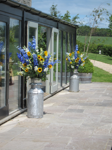Sunflowers, Delphinium, and mixed foliage milk churn displays at Cripps Stone Barn. Floral design by Cotswold Blooms, wedding florist based in Cheltenham.