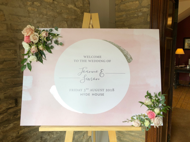 Welcome sign with flower sprays dressing each corner. Floral design by Cotswold Blooms, wedding florist based in Cheltenham.