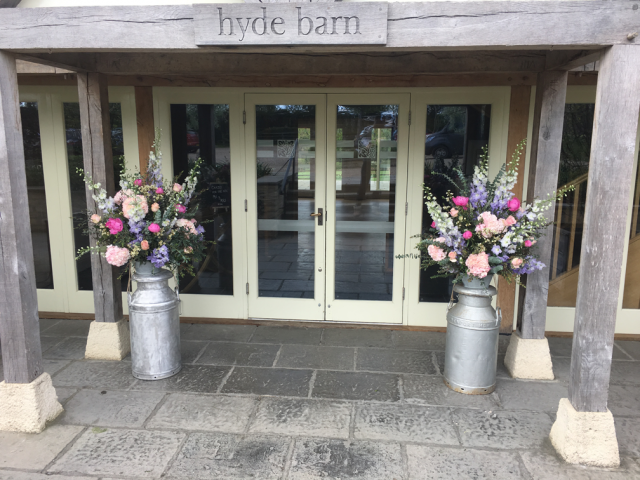 Lilac and pink milk churn display including Delphinium, Peonies and Roses . Floral design by Cotswold Blooms, wedding florist based in Cheltenham.