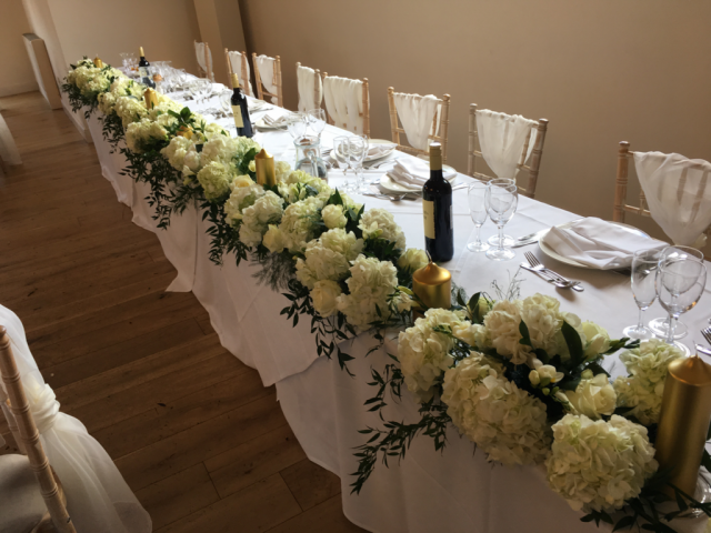 Long table runner in a white and gold colour theme, containing Hydrangeas, Roses and Freesia. Floral design by Cotswold Blooms, wedding florist based in Cheltenham.