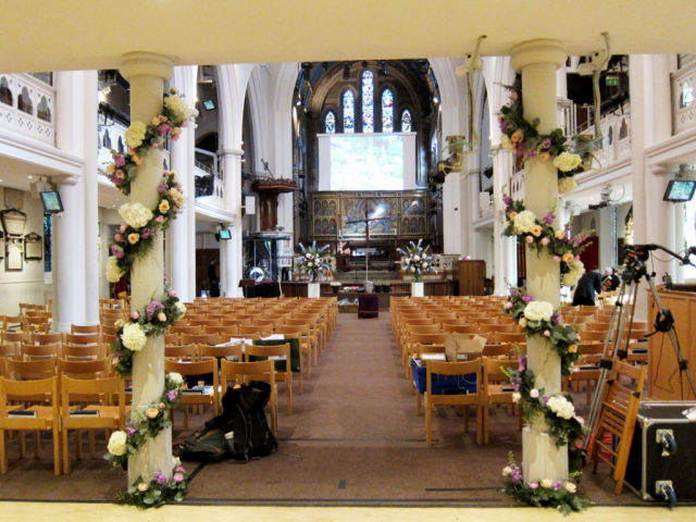 Pillars wrapped with flower garlands and pedestal displays at Holy Trinity Brompton, London. Floral design by Cotswold Blooms, wedding florist based in Cheltenham.