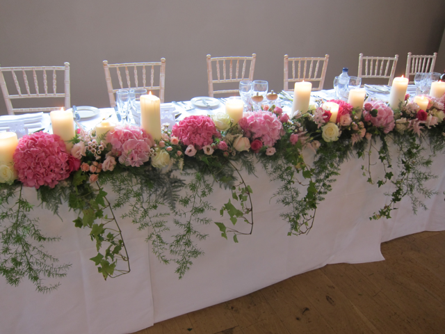 Full length top table display that trails forward including Hydrangea, Roses, Eustoma and candles. Floral design by Cotswold Blooms, wedding florist based in Cheltenham.