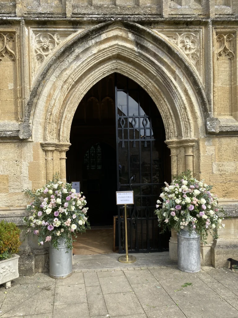Large classic wedding floral installation with milk churns at the church entrance