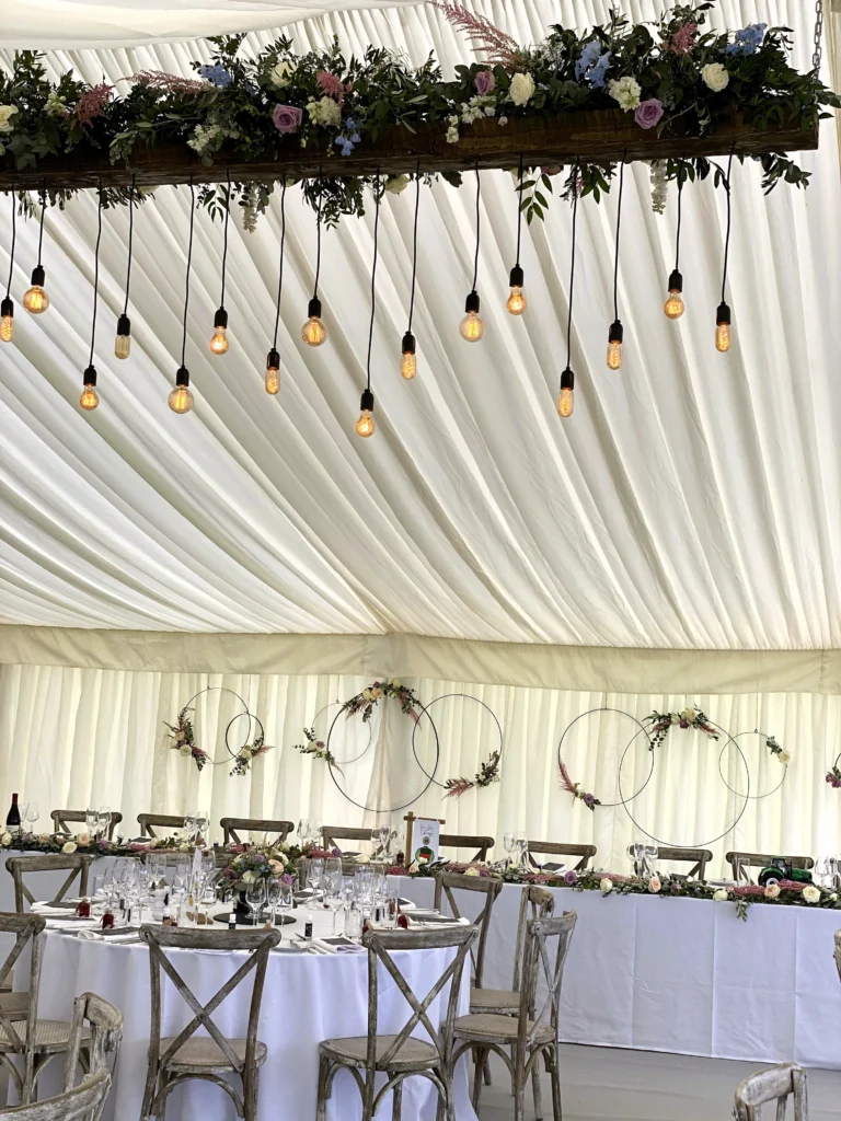 Floral hoop top table backdrop, dressed festoon lights and table centrepieces inside a rustic marquee wedding